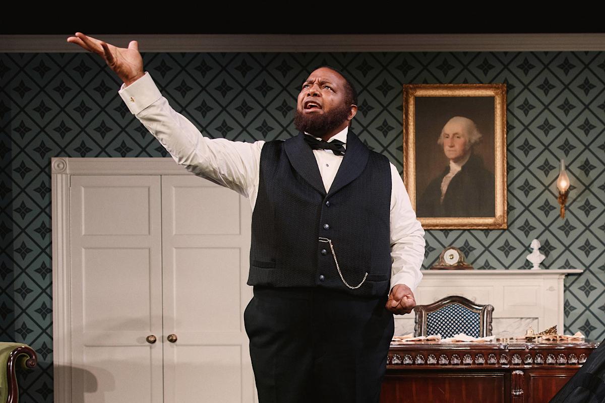 A man in formal attire stands in a vintage room, raising one hand expressively while performing. Behind him is a portrait of George Washington, patterned wallpaper, a desk, and a lit candle.