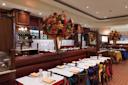 A spacious, elegant restaurant with wooden booths, colorful tablecloths, large floral centerpieces, set wine glasses, and a warm, inviting ambiance. Shelves with bottles and framed art decorate the background.