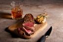 A glass of amber whiskey sits beside a wooden board with sliced medium-rare steak and roasted garlic garnished with rosemary. A knife rests next to the board on a marble surface.