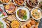 A variety of dishes on a wooden table, including steak with green beans, macaroni and cheese, fries with herbs, pita bread with dip, sliders, salad, wings, and chocolate cake dessert.