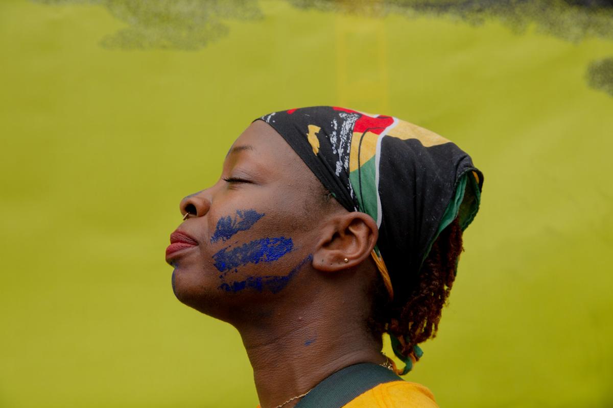 A person in profile with eyes closed, wearing a colorful headscarf and yellow shirt. Blue paint marks are visible on their cheek. The background is bright yellow.