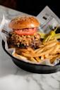 A sesame seed burger bun filled with a beef patty, crispy fried onions, and tomato slices, served with golden French fries, pickles, and a yellow pepper in a black basket lined with paper.
