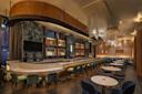 Sleek modern bar interior with a marble counter and high stools on the left, and tables with chairs on the right. The space features dim lighting, wooden accents, hanging light fixtures, and a wall-mounted display of bottles.