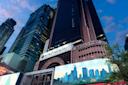 A tall Hyatt Regency hotel building stands among skyscrapers in a city at dusk, with a Broadway sign and a billboard featuring a city skyline in the foreground. The sky is partly cloudy.