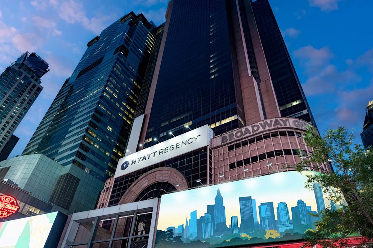 A tall Hyatt Regency hotel building stands among skyscrapers in a city at dusk, with a Broadway sign and a billboard featuring a city skyline in the foreground. The sky is partly cloudy.