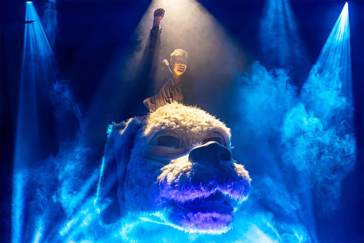A person with a raised fist rides a large, white, furry creature’s head, surrounded by blue stage lights and mist, creating a dramatic and fantastical atmosphere.