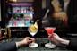 Two people hold cocktails at a bar. One drink is in a stemmed glass with an orange slice and rosemary garnish; the other is a pink drink in a martini glass. The background features a dimly lit bar and a blurred painting.