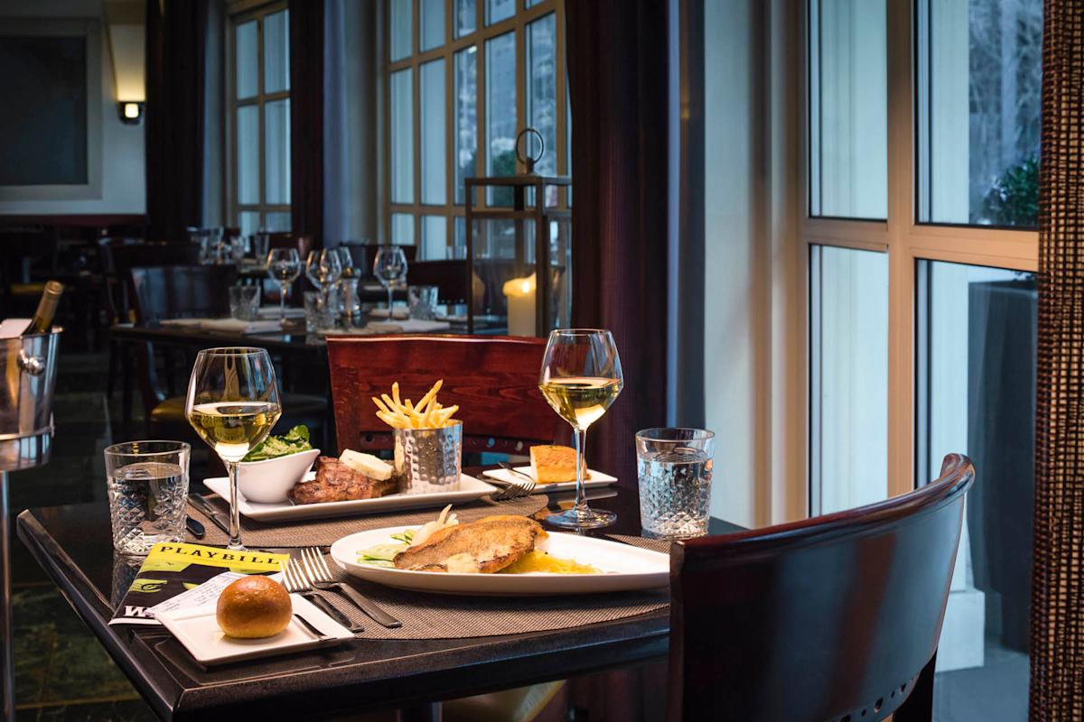 A cozy restaurant table set for two, featuring plates with meat, vegetables, and fries. Two glasses of white wine accompany the meal. A Playbill and a roll are on the table. Large windows provide ambient natural light.