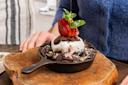 A small cast iron skillet filled with a chocolate dessert topped with a scoop of vanilla ice cream, chocolate sauce, sliced strawberries, and mint leaves. It's placed on a wooden board, with a person sitting nearby.