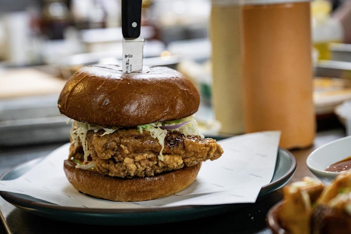 A fried chicken sandwich with coleslaw on a brioche bun sits on a plate, with a knife stabbed through the top. Blurred kitchen items and a bottle of sauce are visible in the background.
