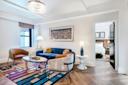 Modern hotel suite living area with a blue velvet sofa, two brown chairs, a round coffee table, a colorful rug, a cityscape painting, and a chandelier. Bedroom with a bed visible through open doorway.
