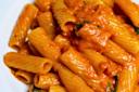 Close-up of rigatoni pasta covered in a creamy tomato sauce with pieces of basil. The dish is served on a white plate, showcasing the sauce's rich texture and the pasta's ridged surface.