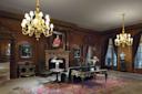 Ornate room with wood paneling, chandeliers, and antique furniture. A large rug covers the floor, portraits hang on the walls, and a marble fireplace with a painting above completes the elegant, classical decor.