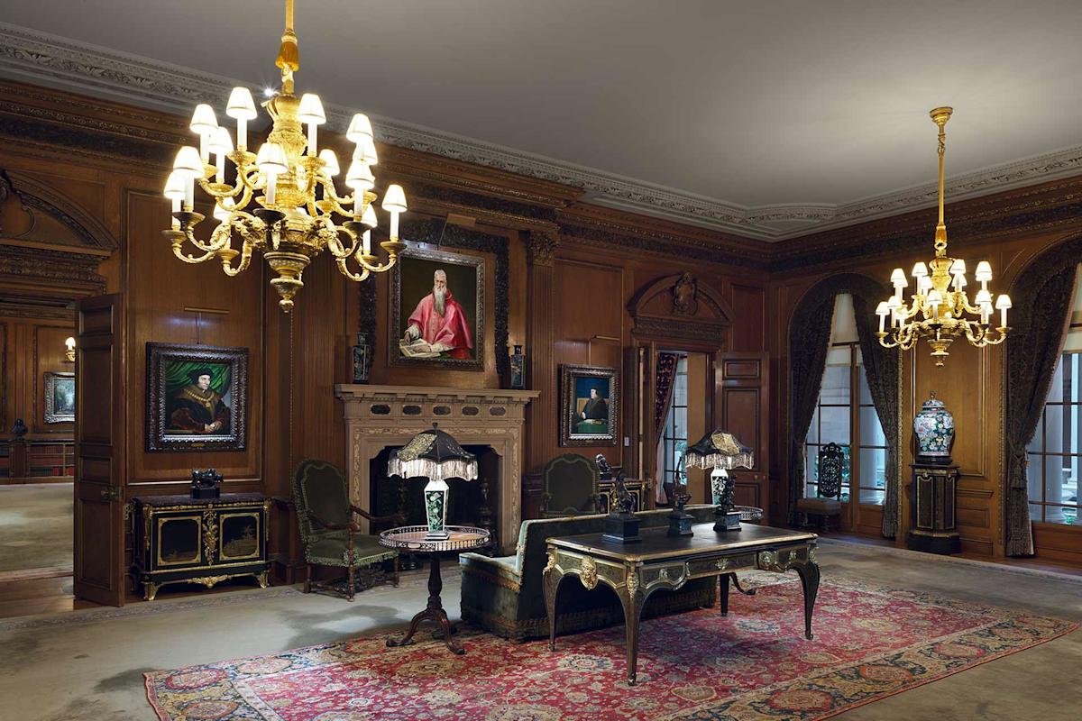 Ornate room with wood paneling, chandeliers, and antique furniture. A large rug covers the floor, portraits hang on the walls, and a marble fireplace with a painting above completes the elegant, classical decor.
