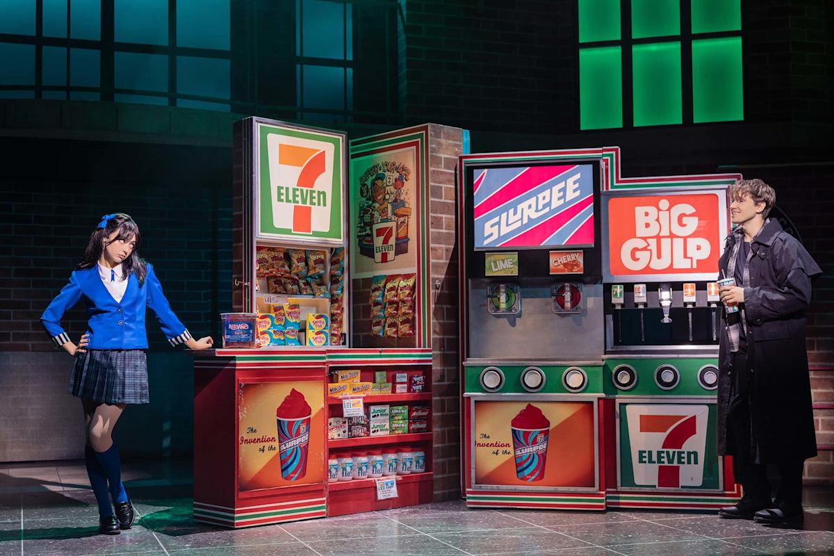 Two people in costumes stand by colorful 7-Eleven snack and drink machines onstage. The left person wears a blue school uniform, while the right person in dark clothes holds a drink, both in a theatrical setting.
