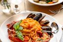 A plate of seafood pasta features mussels, lobster, and tagliatelle noodles garnished with fresh basil. A spoon and fork rest on the side. In the background, a glass of water and another seafood dish can be seen on the table.
