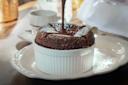 A chocolate soufflé in a white ramekin is being topped with chocolate sauce, set on a decorative white plate with a doily, next to a cup and saucer on a wooden table in natural light.