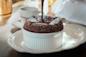 A chocolate soufflé in a white ramekin is being topped with chocolate sauce, set on a decorative white plate with a doily, next to a cup and saucer on a wooden table in natural light.