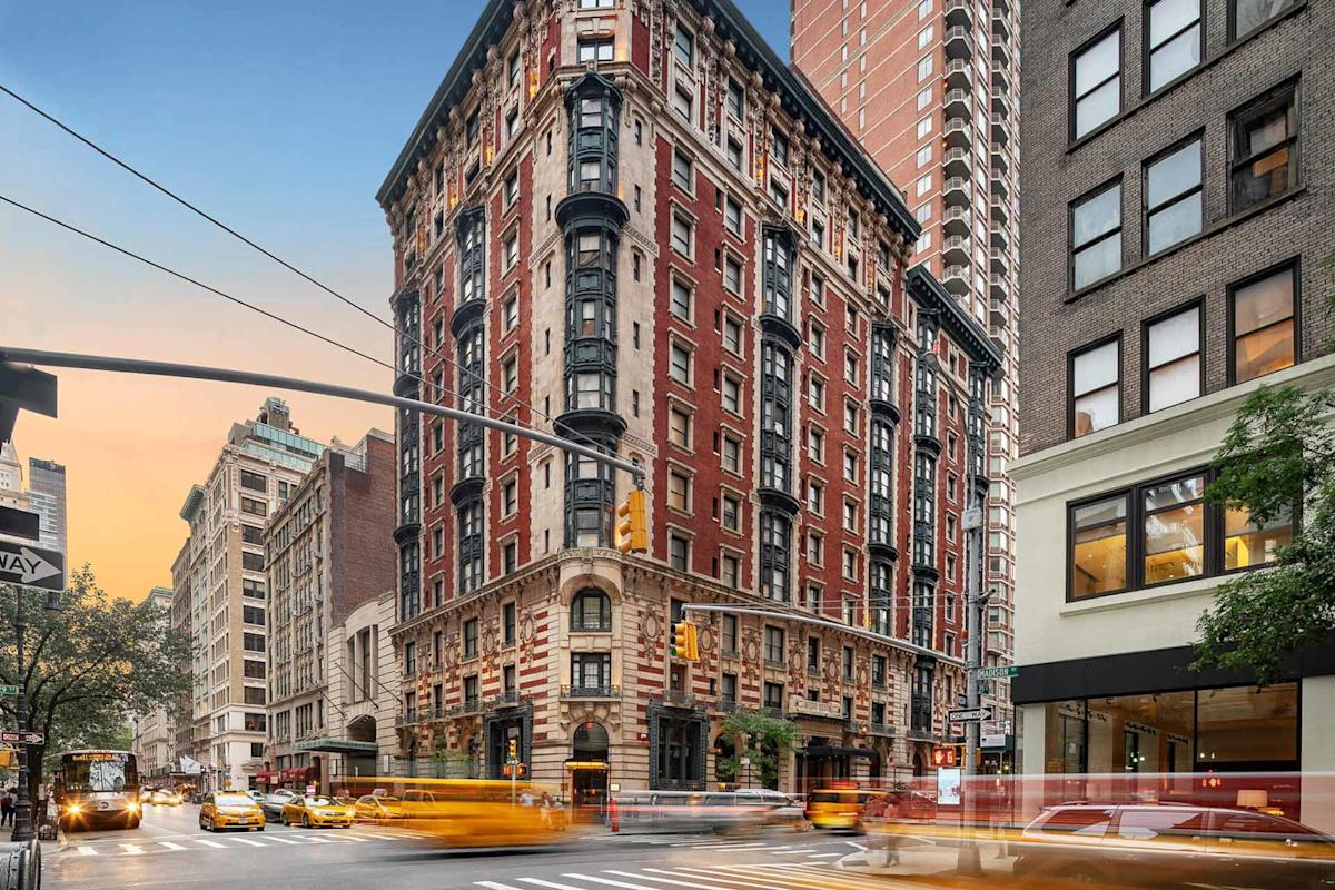 A busy New York City street corner at sunset with a historic red-brick apartment building, yellow taxis, and blurred motion from traffic and people.