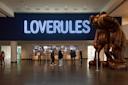 People stand in a museum lobby under a large neon sign that reads "LOVERULES." To the right, there's a large brown sculpture depicting two humanoid figures leaning forward. In the background, a counter with people and displays is visible.
