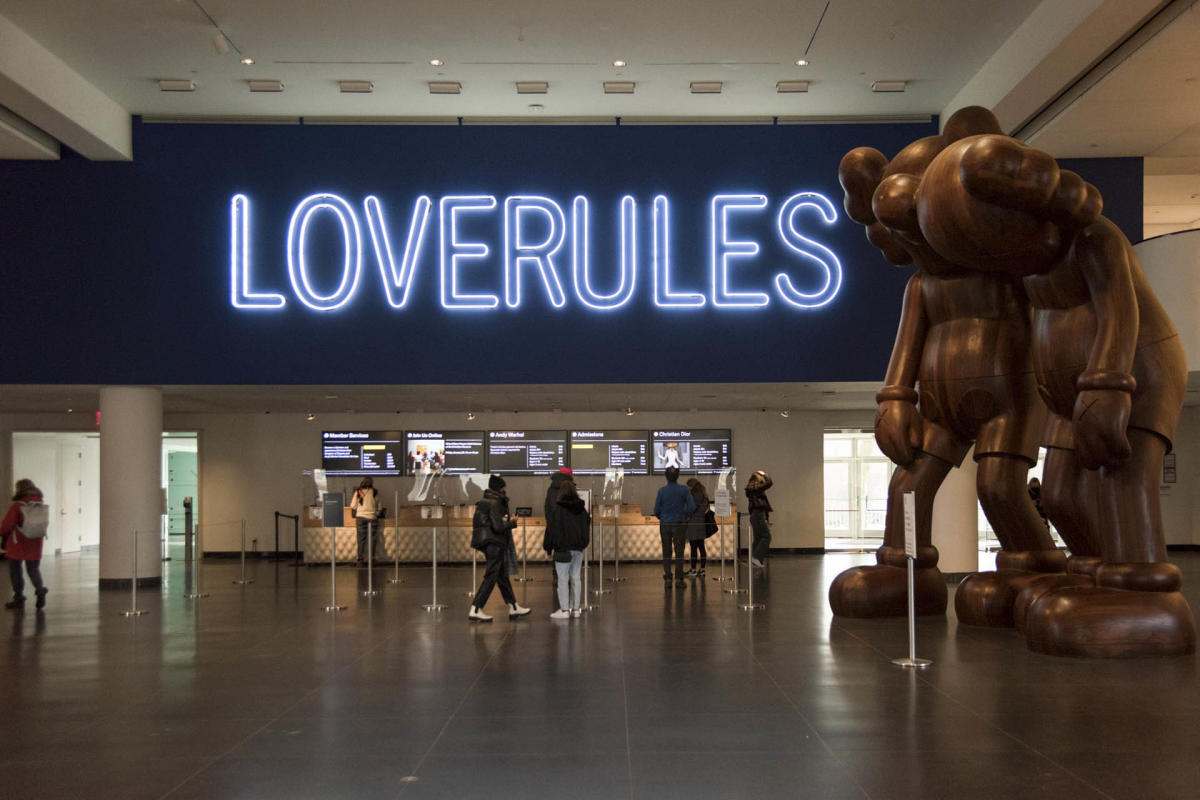 People stand in a museum lobby under a large neon sign that reads "LOVERULES." To the right, there's a large brown sculpture depicting two humanoid figures leaning forward. In the background, a counter with people and displays is visible.