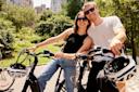 A smiling couple wearing sunglasses poses with their bicycles in a sunny park, surrounded by green trees and tall city buildings in the background.