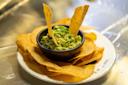 A black bowl of guacamole is surrounded by golden tortilla chips on a white plate. Three chips are upright in the guacamole. The background is metallic and blurred.