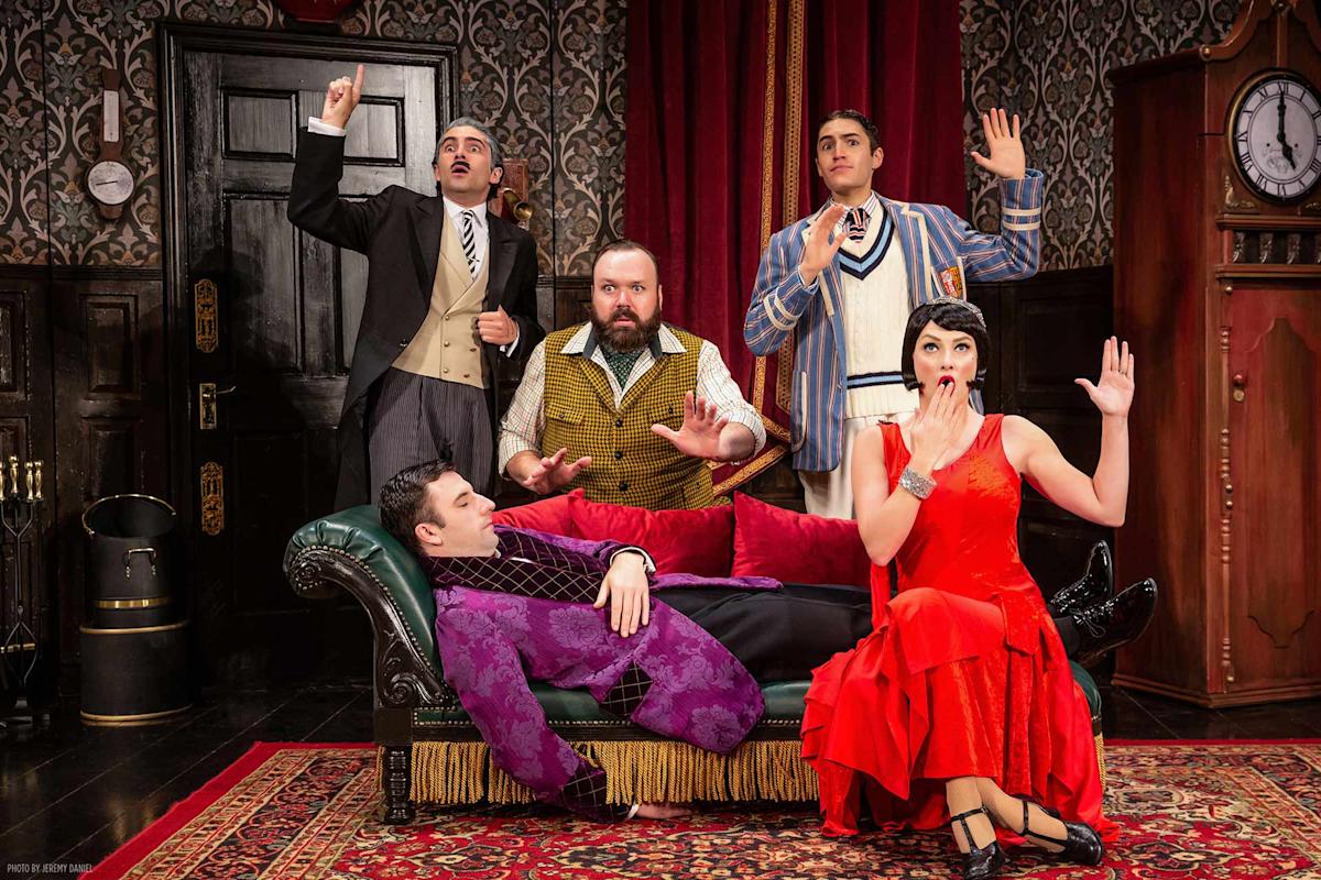 Five actors in vintage costumes pose dramatically on a stage set resembling an old-fashioned living room, with one man lying on a green sofa and the others standing or sitting and raising their hands as if startled or surprised.