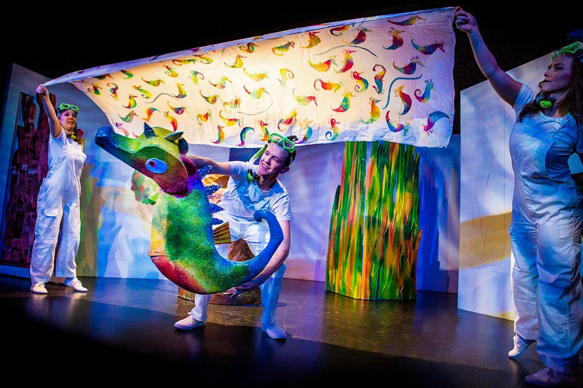Three performers in white costumes hold a colorful sea creature puppet and a large, decorated fabric backdrop on stage, with bright, playful lighting and props.