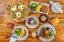 A wooden table set with a variety of brunch dishes, including a breakfast pizza with eggs, steak, eggs Benedict with potatoes, an omelet, salad, toast, coffee, and two colorful juices.