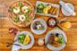 A wooden table set with a variety of brunch dishes, including a breakfast pizza with eggs, steak, eggs Benedict with potatoes, an omelet, salad, toast, coffee, and two colorful juices.