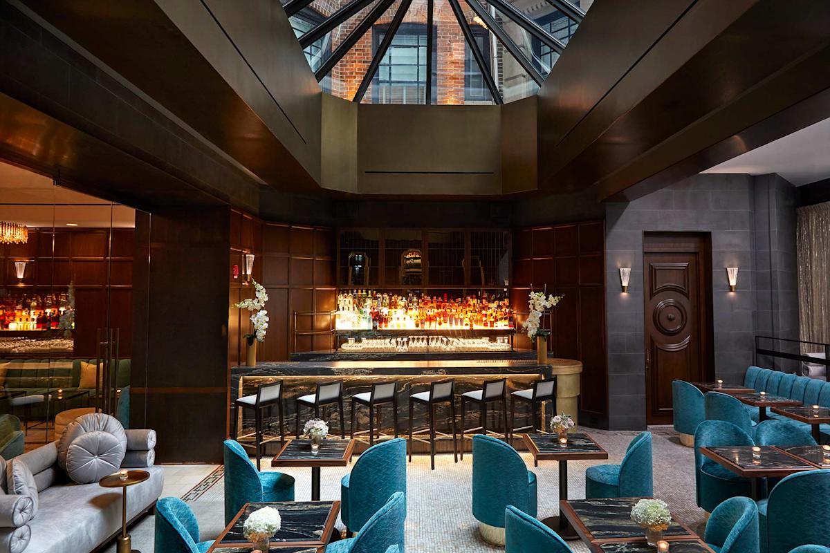 A modern, elegant bar with a glass ceiling, dark wood paneling, a large backlit bar, and blue velvet chairs around small tables. Natural light streams in, highlighting the stylish decor and floral arrangements.