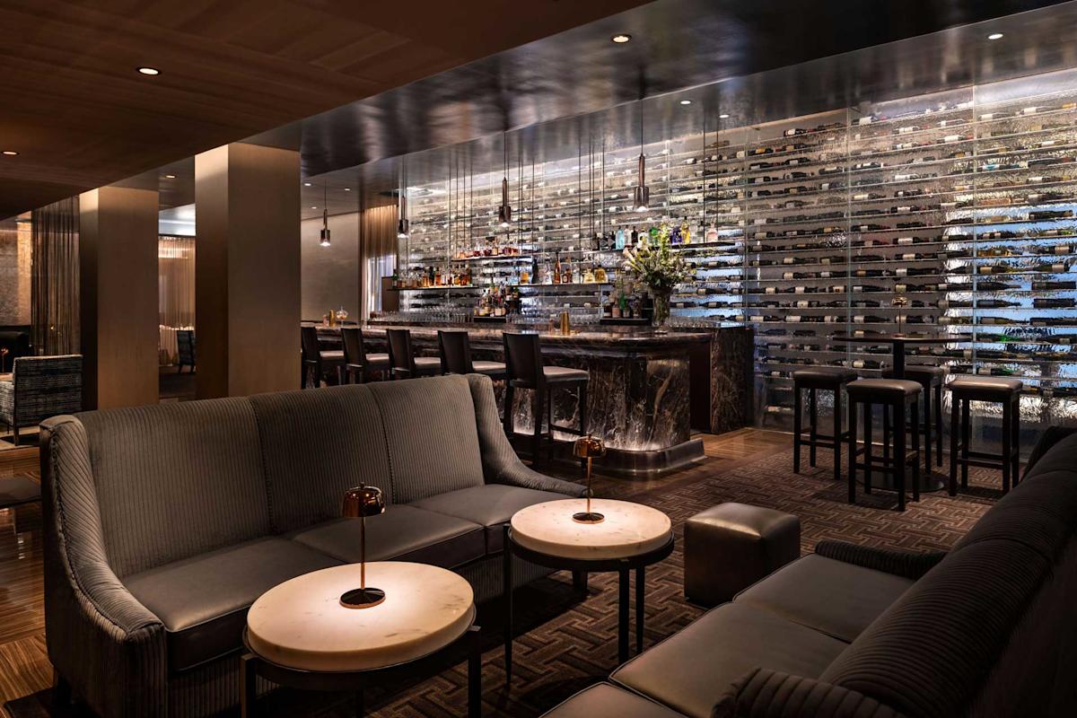 The martini lounge bar interior featuring plush seating arrangements around small tables. A sleek bar counter with high stools is in the background, adorned with various bottles and flowers. The ambiance is luxurious and inviting.