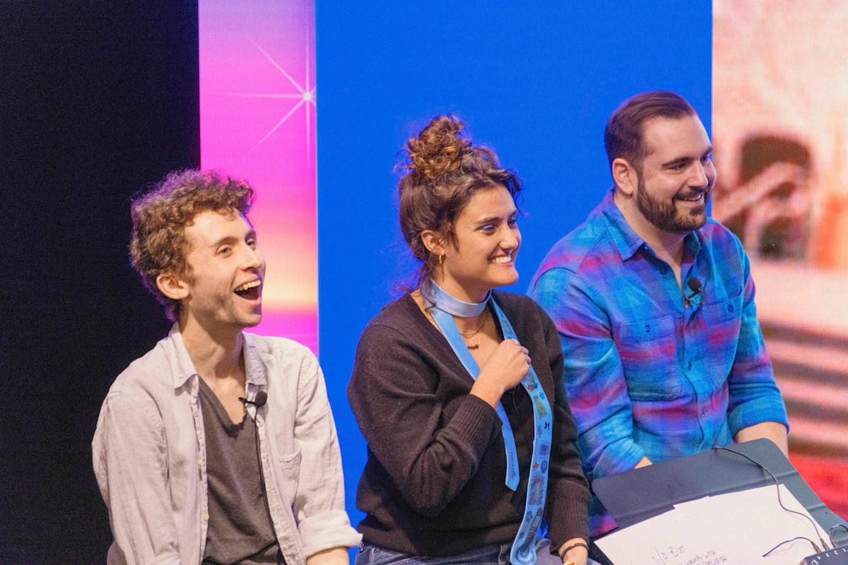 Three people are sitting and smiling on a stage. They appear to be enjoying a presentation.