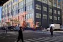 People cross a city street at a corner with a large industrial-style building featuring colorful geometric wall art. Parked cars and a white truck line the streets under sunny skies.