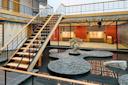 A modern indoor space features a wooden staircase over a Zen garden with pebbles, rocks, and a small tree, surrounded by water and glass walls with a view of an open, well-lit area.