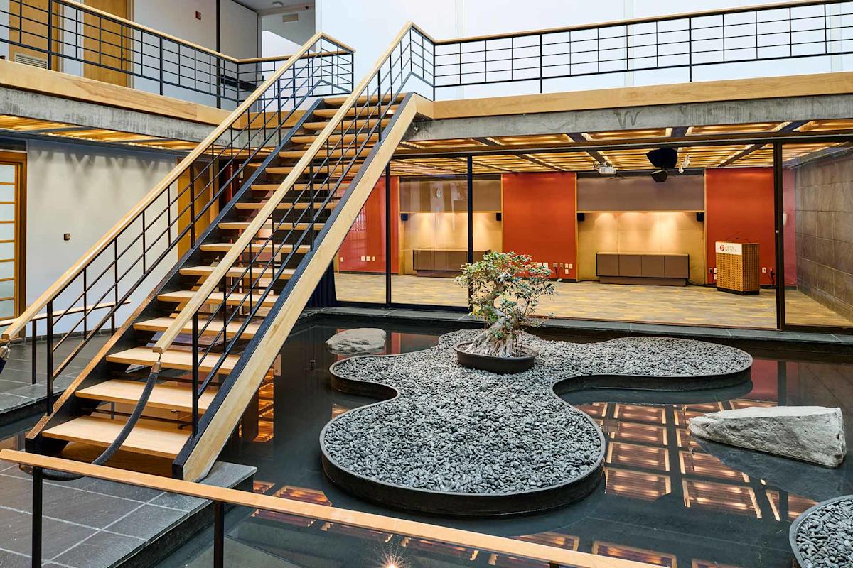 A modern indoor space features a wooden staircase over a Zen garden with pebbles, rocks, and a small tree, surrounded by water and glass walls with a view of an open, well-lit area.