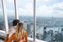 A couple embraces while gazing out a large window at a sprawling cityscape, featuring a river and tall buildings. The scene is viewed from a high vantage point on a clear day.