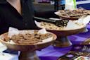 Three wooden bowls on a purple table, each filled with different chocolate treats: chocolate squares, powdered candies, and chocolate-covered pretzels with sprinkles. A person in a black sweater stands behind the bowls.