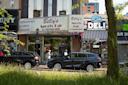 Street view of shops including Billy’s Sports Bar Restaurant and Grand Slam Deli, with cars parked in front, people walking on the sidewalk, and a grassy area and tree in the foreground.