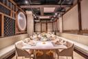 Elegant restaurant dining area with modern decor, a long table set with beige napkins, white flowers as centerpieces, light-colored chairs, and cushioned bench seating along the walls under soft lighting.
