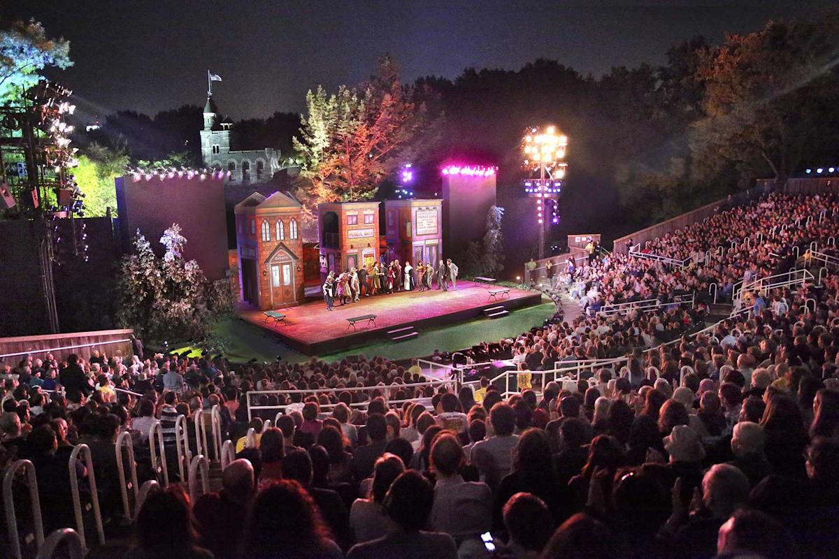 An outdoor theater at night with a full audience watching a stage play set in front of building facades, surrounded by trees and illuminated by colorful stage lights.