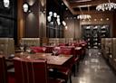 Modern restaurant interior with red leather chairs, wooden tables set with wine glasses and silverware, beige upholstered booths, wine rack, large windows, and decorative pendant lights. The space is empty and well-lit.