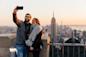 A couple taking a selfie on a rooftop with a city skyline in the background at sunset. They are smiling, and the Empire State Building is visible in the distance. The woman is wearing a scarf and coat, and the man is wearing glasses and a jacket.