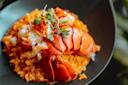 A dish featuring a cooked lobster tail served on a bed of golden saffron rice, garnished with microgreens. The vibrant colors contrast with the dark plate, creating an elegant presentation.