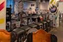 A row of green cushioned bar stools lines a railing above a bookstore filled with shelves and artwork; colorful, painted columns with abstract designs are visible throughout the space.