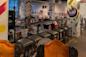 A row of green cushioned bar stools lines a railing above a bookstore filled with shelves and artwork; colorful, painted columns with abstract designs are visible throughout the space.