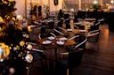 A warmly lit outdoor dining area featuring neatly arranged tables with chairs. Each table is set with white napkins and glassware. A decorated Christmas tree with silver ornaments is visible in the foreground.