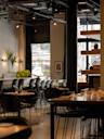 A modern, stylish restaurant interior with black chairs and wooden tables, set with glasses and small vases. Large windows let in natural light, and decorative plants and bananas are visible in the background.