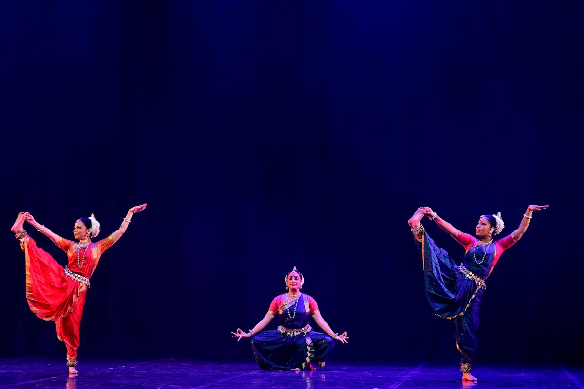 Three dancers in colorful traditional costumes perform on stage; two stand with one leg raised and arms extended, while the center dancer sits cross-legged, all against a deep blue background.